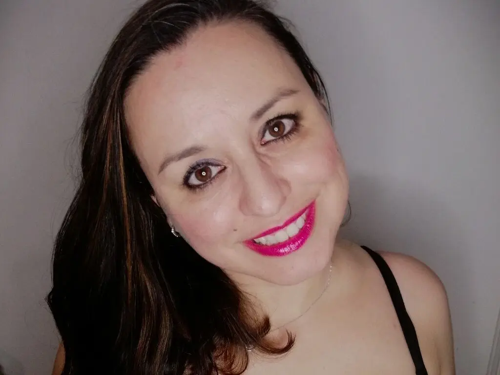 Close-up selfie of a young woman with slicked-back hair and red lipstick, wearing an orange top in dappled sunlight outdoors.
