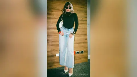 Stylish woman in black turtleneck and light wash wide-leg jeans posing against a wooden wall.