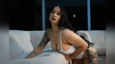 JuliahLewis poses in a white plunging bodysuit while leaning against a white tufted sofa.