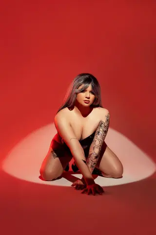 Sensual woman with arm tattoos posing in black lingerie against a dramatic red studio background.