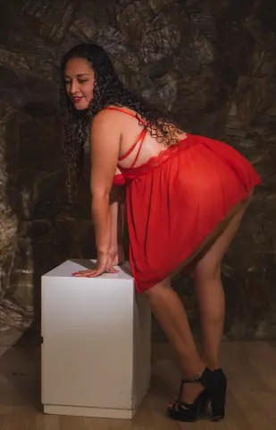Smiling woman in a red slip dress leaning on a white pedestal against a dark stone wall background.