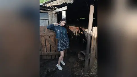 Young woman in a denim jacket and plaid skirt posing in a rustic wooden barn setting.