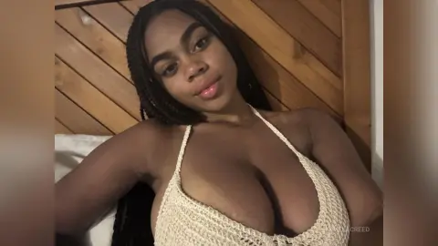 Young Black woman with long braids wearing a white crochet bikini top relaxing in a cozy bedroom setting.