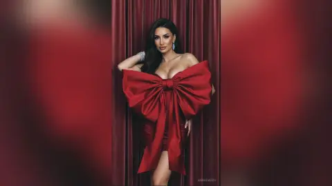 AnnaKarev poses in a red dress with a massive bow in front of red curtains.
