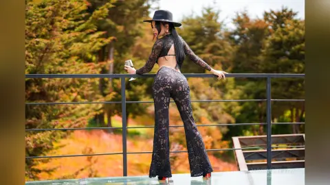 Stylish woman in black lace jumpsuit and wide-brim hat posing on a balcony overlooking autumn trees.