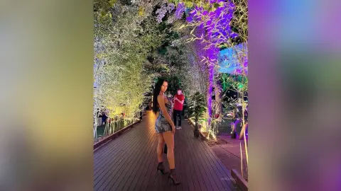 NicoleBella stands on a wooden walkway wearing a blue denim romper and dark heels, surrounded by lush green trees and purple lighting.