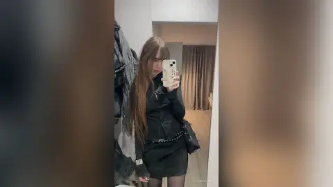 Young woman with long hair in a black jacket and skirt taking a mirror selfie in a modern room.