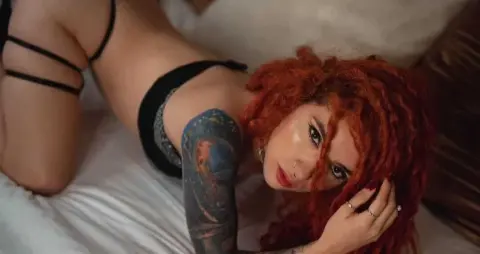 Redheaded woman with tattoos in black lingerie relaxing on a white bed in a moody, intimate setting.