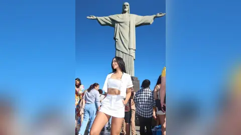 Alison stands outdoors in a white crop top and matching white shorts against a clear blue sky with a large statue behind her.
