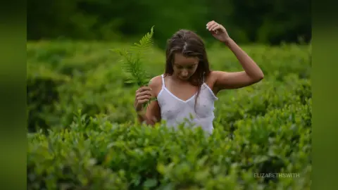 Young woman in a white camisole holding a green fern in a lush, dreamy forest setting.