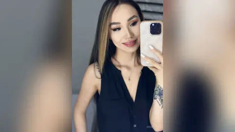 Smiling young woman in a black sleeveless top taking a mirror selfie in a bright indoor setting.