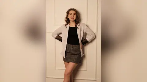 Smiling young woman in a white blazer and black top posing against a white door in a bright indoor setting.