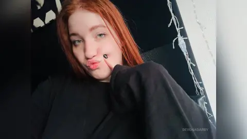 Redheaded woman in a black sweatshirt making a playful pouty face in a dimly lit room with fairy lights.