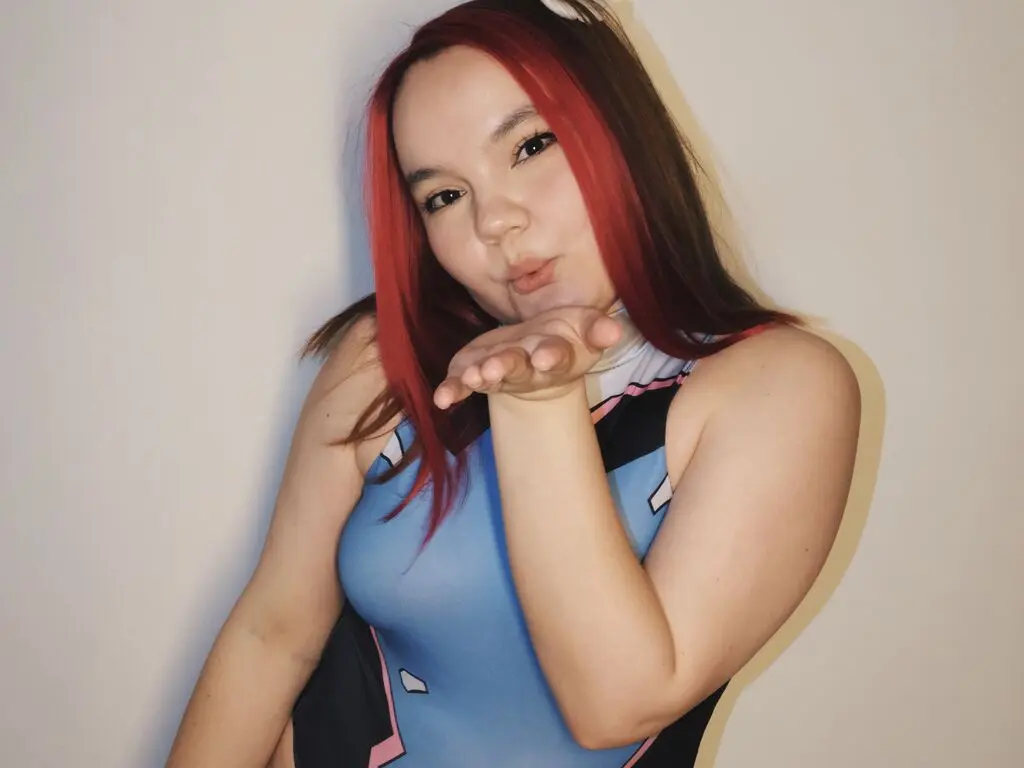 Red-haired woman in a blue and black sleeveless top blowing a kiss against a plain white wall.