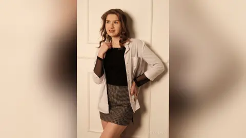 Young woman in a black top and plaid skirt leaning against a white door in a soft, moody indoor setting.