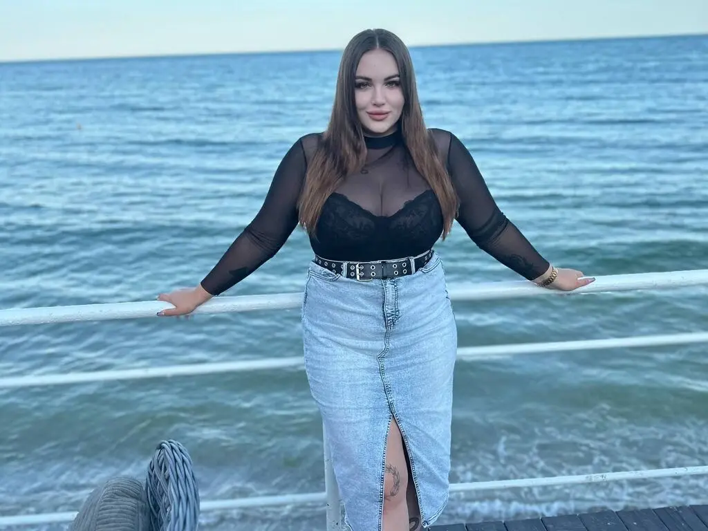 Young woman in a black sheer top and denim skirt posing by the ocean at sunset.