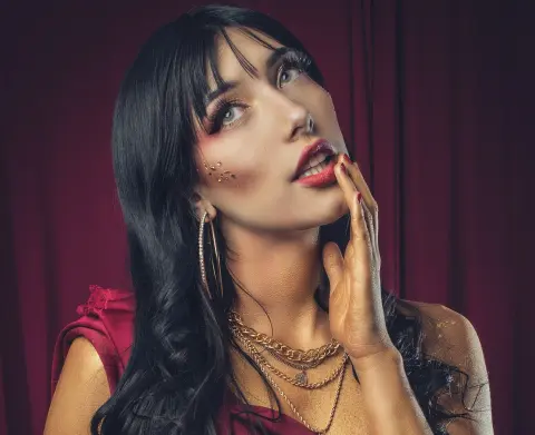 Glamorous woman with dark hair and gold glitter makeup wearing red outfit and gold jewelry against a velvet backdrop.