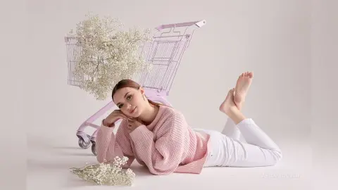 Woman in pink knit sweater and white pants relaxing next to a shopping cart filled with white flowers.