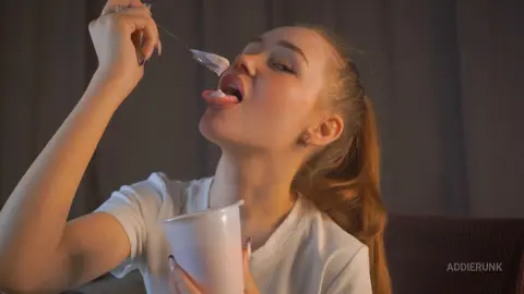 AddieRunk eats white cream from a spoon, tilting her head back with her mouth open and tongue out while wearing a white t-shirt.