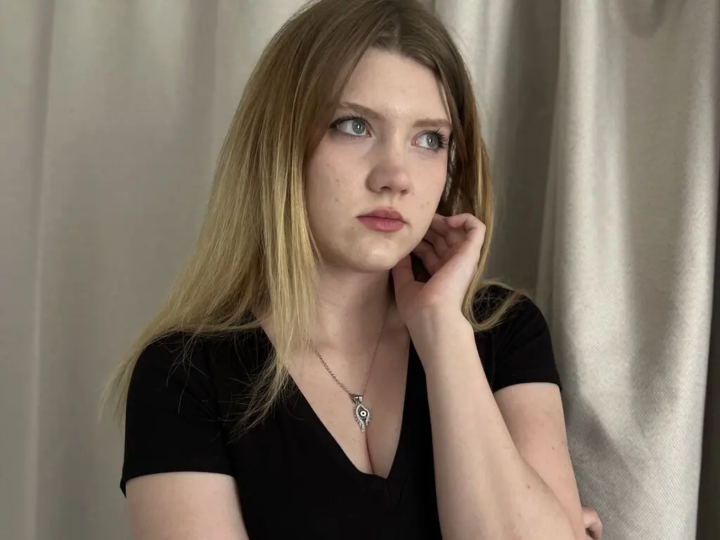 GemmaCiliberto looks off to the side with long blonde hair, wearing a black v-neck t-shirt and an evil eye pendant against a white curtain background.