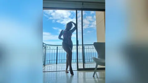 Young woman in a floral swimsuit posing on a sunny balcony overlooking the blue ocean.