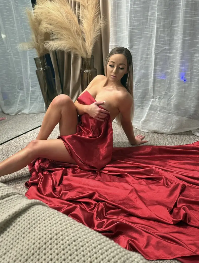 JessicaBennett poses naked from the waist up, wearing a red silk slip that drapes over her legs while she sits on red satin bedding.