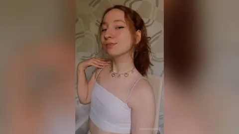MadgeRemerez poses in a white camisole, showing her neck, shoulders, and light brown hair against a patterned wall background.