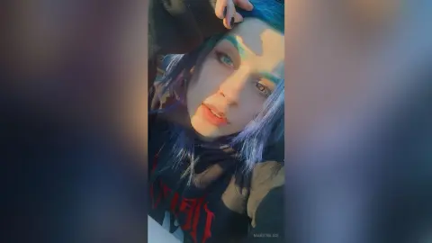 Young woman with blue hair wearing a black graphic hoodie posing in warm sunlight.