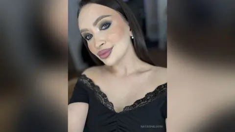 NatashaLeoni poses with long dark hair and heavy makeup, wearing a black lace off-the-shoulder top.