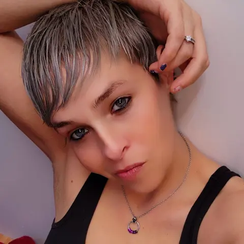 Woman with short grey pixie cut wearing a black tank top and silver necklace in a soft indoor setting.