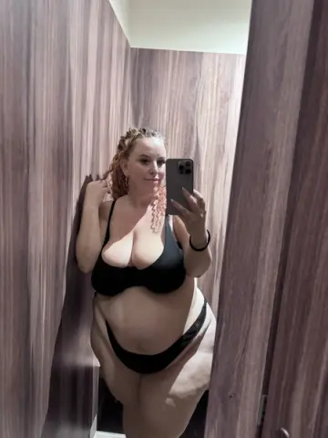 Curvy blonde woman in black lingerie taking a mirror selfie in a modern wood-paneled bathroom.
