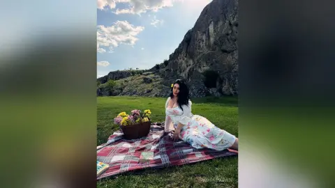 Woman in a white floral dress sitting on a picnic blanket near a mountain under a sunny sky.