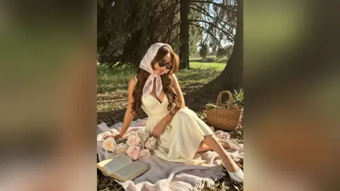 Stylish woman in a white sundress and headscarf relaxing on a picnic blanket in a sunny park setting.