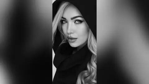 Anelise poses with a direct gaze, wearing a black hooded top that frames her blonde hair and heavy eye makeup.