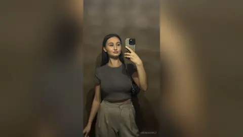 Young woman in a grey crop top and beige trousers taking a mirror selfie in a moody, dimly lit room.