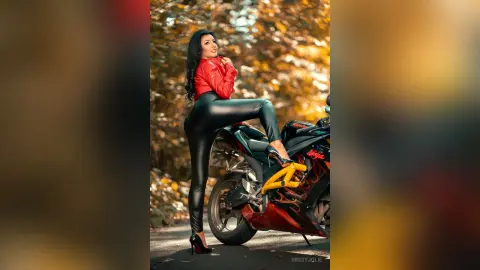 MissyJolie poses nakedly in black leggings and a red top while sitting on a motorcycle against a background of autumn leaves.