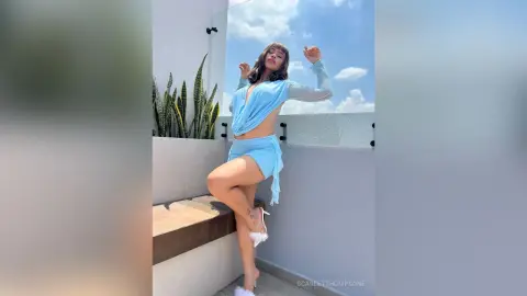 Young woman in a light blue two-piece outfit posing on a sunny balcony with a bright sky background.