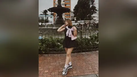 Young woman in a black summer dress and white sneakers posing by a city fountain on a sunny day.