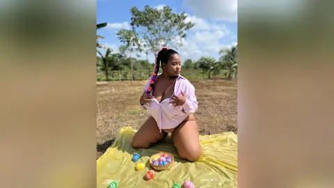 Curvy woman in a light pink shirt sitting on a yellow blanket outdoors near colorful Easter eggs.