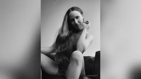 Elegant woman in a dark sleeveless dress posing thoughtfully in a minimalist indoor setting, black and white portrait.