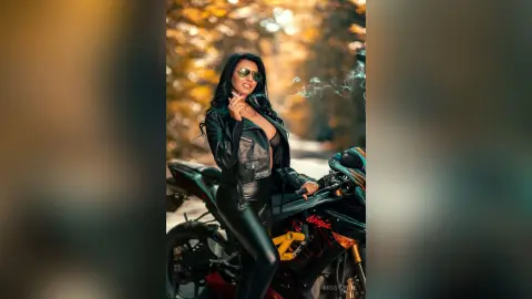 MissyJolie wears a black leather jacket and tight leggings while posing next to a black motorcycle outdoors.