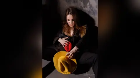 Moody portrait of a woman in a black outfit sitting in shadows with a bright yellow hat.