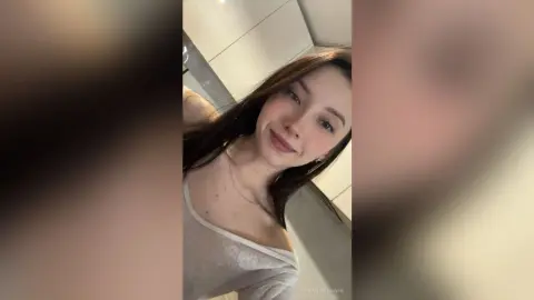 Smiling young woman with long brown hair wearing a white casual top in a bright indoor setting.