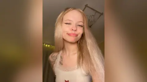LucyLoy winks at the camera while wearing a white lace camisole, showing off her long blonde hair and arm tattoo.