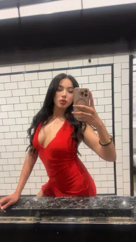 Young woman in a red cocktail dress taking a mirror selfie in a modern white tiled bathroom.