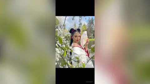 IngridSaint poses in a white and red outfit holding a folding fan amidst blooming white flowers in a park.