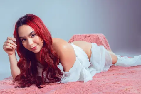 Smiling woman with long red hair in a white flowing dress lying on a pink faux fur rug in a bright studio.