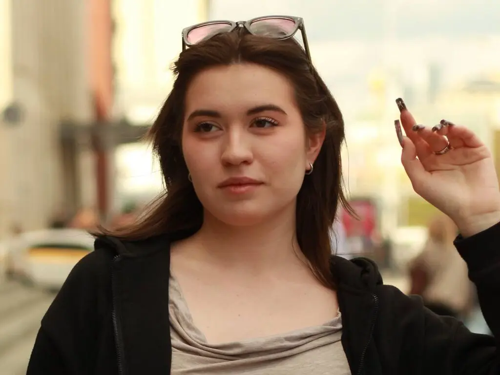 Young woman in a black hoodie and beige top looking thoughtful in an urban outdoor setting.