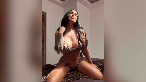 Lexi poses in a patterned bikini, showing off her tattoos and long dark hair while sitting on a leopard print bed.