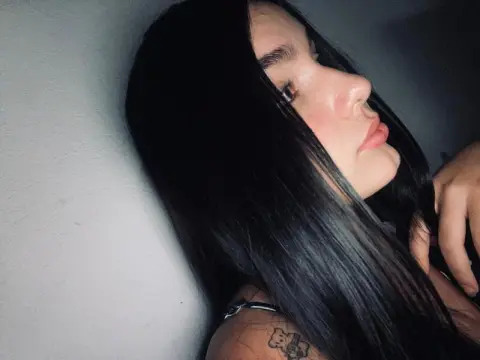 Close-up portrait of a young woman with long dark hair posing against a white wall in soft indoor lighting.